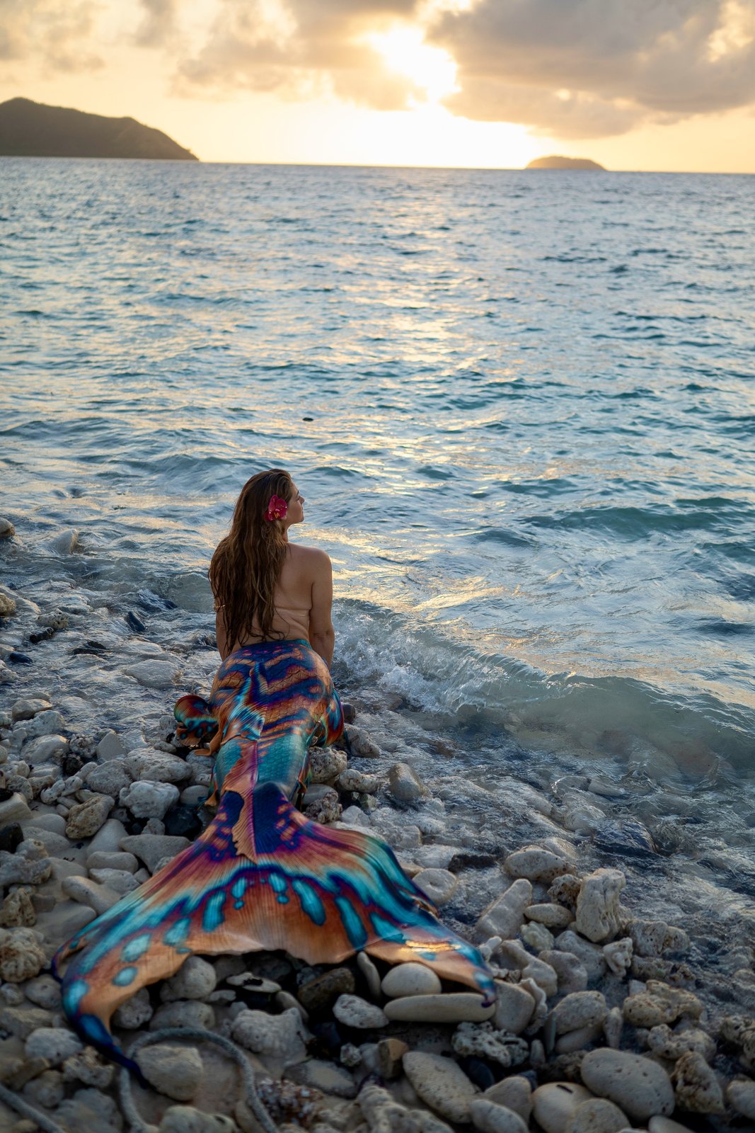 Brandee Anthony, the Vero Beach Mermaid, resting on a rocky shore at sunset in a rainbow Finfolk mermaid tail